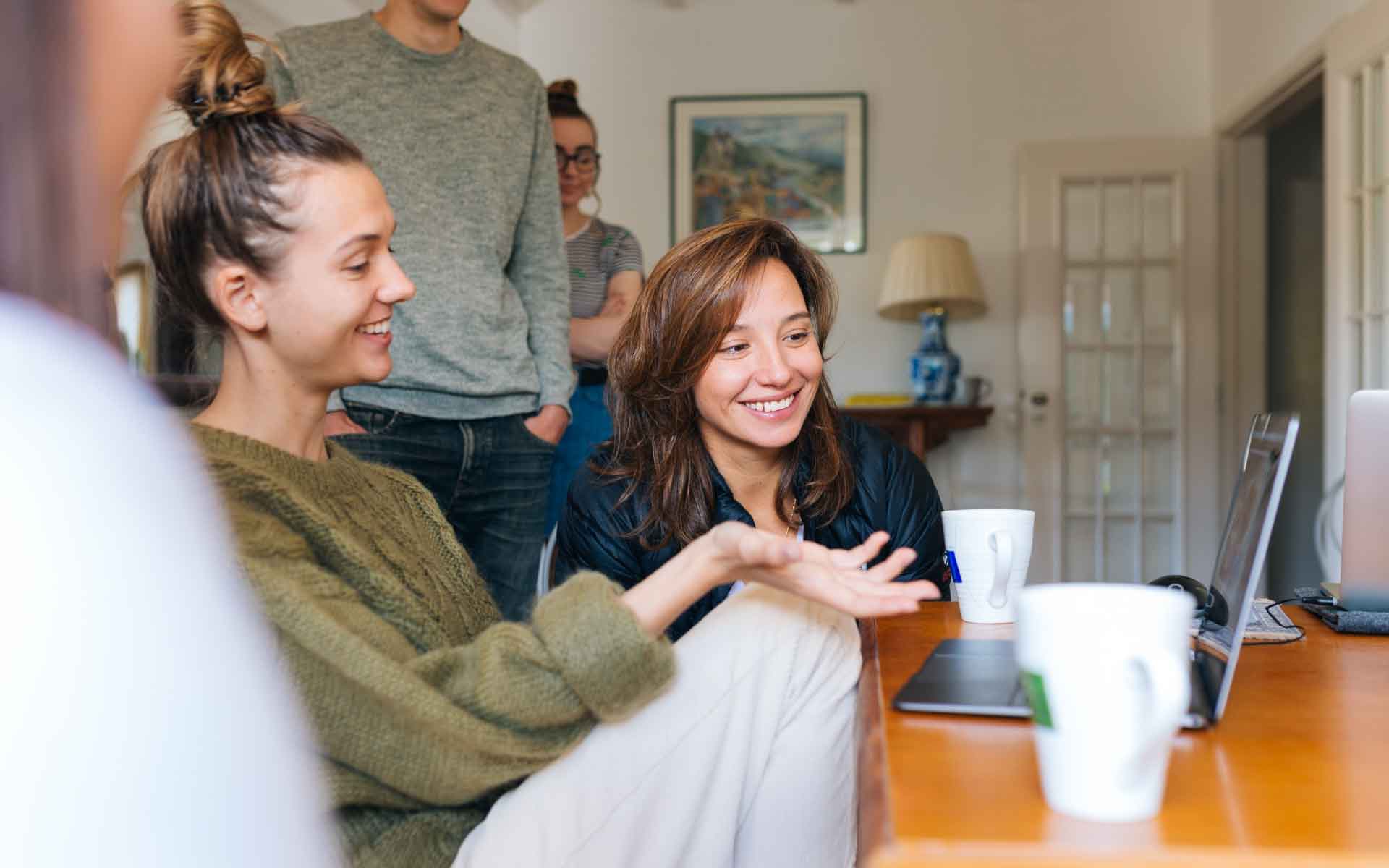Group of people engaging in discussion around a laptop, with a focus on two women smiling and sharing ideas, reflecting collaboration and communication in a home setting.